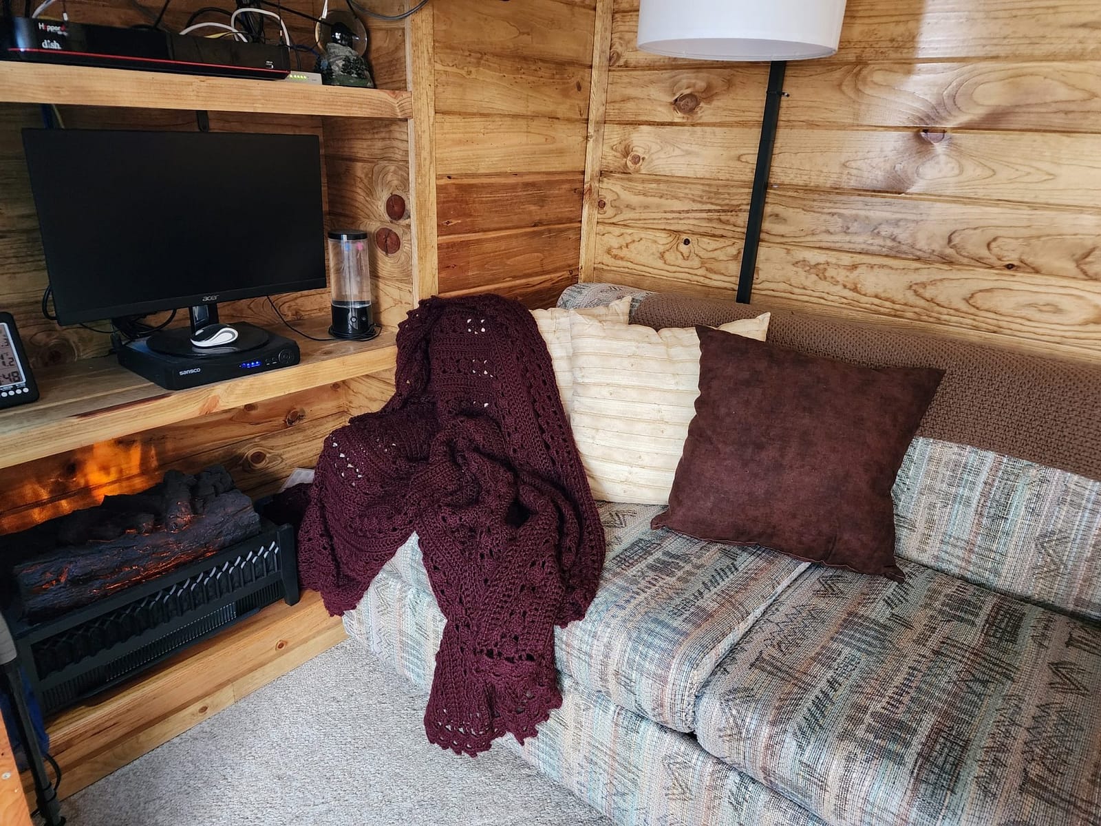 Couch with pillows and afghan on it, a lamp behind it and an electric fireplace next to it